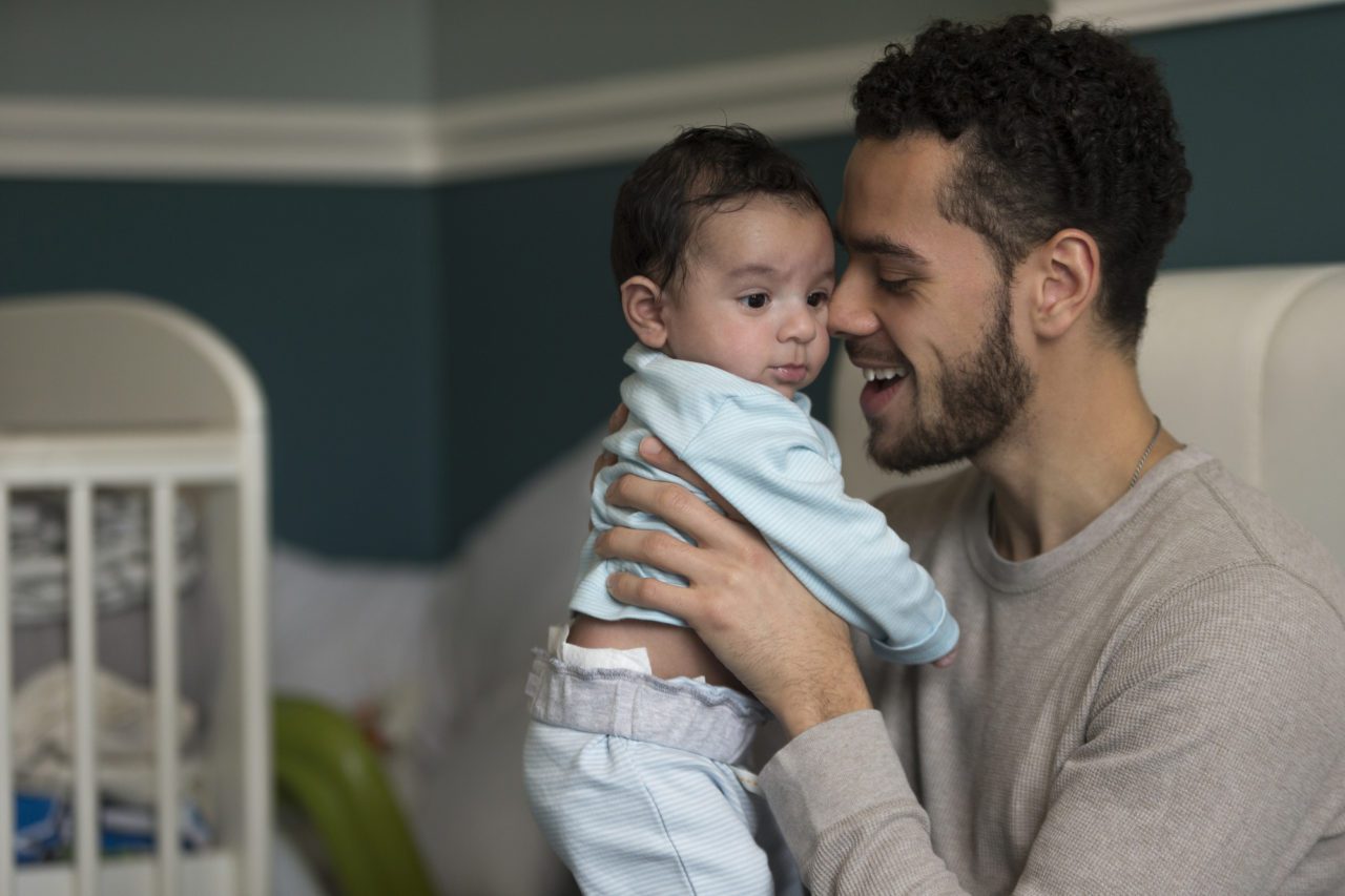 Making Parenting Time Schedules Manageable - Young father sitting on his bed, holding his baby son. He has his nose pressed to his son's cheek.